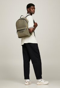 Zaino verde oliva con parte superiore arrotondata, tasca frontale con zip e marchio. Indossato da una persona con una camicia crema e pantaloni scuri.