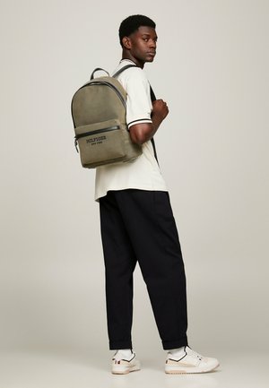 Zaino verde oliva con parte superiore arrotondata, tasca frontale con zip e marchio. Indossato da una persona con una camicia crema e pantaloni scuri.