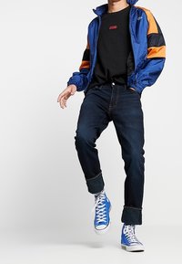 Blå och orange vindjacka med dragkedja, svart t-shirt med röd logga, mörka jeans och blå högsula sneakers.