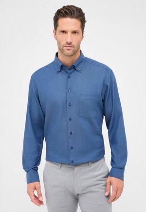 Camicia azzurra a maniche lunghe con finitura testurizzata, taschino a sinistra e bottoni coordinati. Indossata con pantaloni grigi chiari.