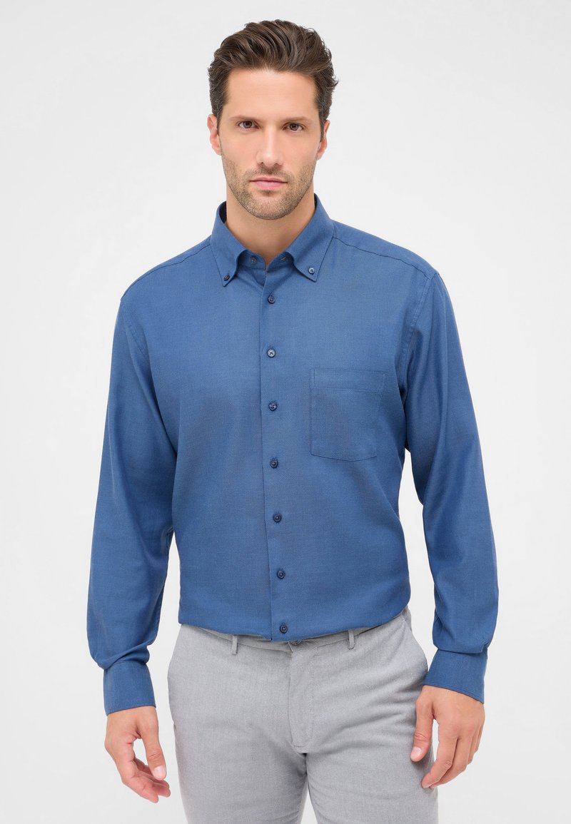 Camicia azzurra a maniche lunghe con finitura testurizzata, taschino a sinistra e bottoni coordinati. Indossata con pantaloni grigi chiari.