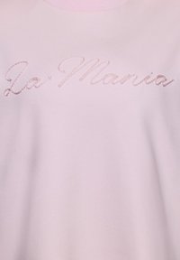 Šviesiai rožinė audinio medžiaga su kursyvu siuvinėtu tekstu „La Mania“ kiek tamsesnės rožinės spalvos siūlais krūtinės srityje.