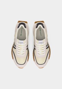 Krém és bézs színű Lacoste tornacipőpár fehér fűzőkkel, fekete oldalsávokkal és barna texturált talpakkal, felülről nézve.