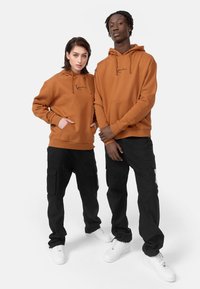 Dos personas llevan sudaderas a juego de color óxido con bolsillos frontales y cordones, combinadas con pantalones cargo negros y zapatillas blancas.