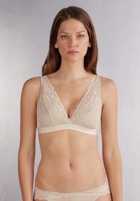 Beige spets-bra med djup V-hals, bred spetskant och mjuk, stretchig underband. Slät yta med intrikata blommönster.