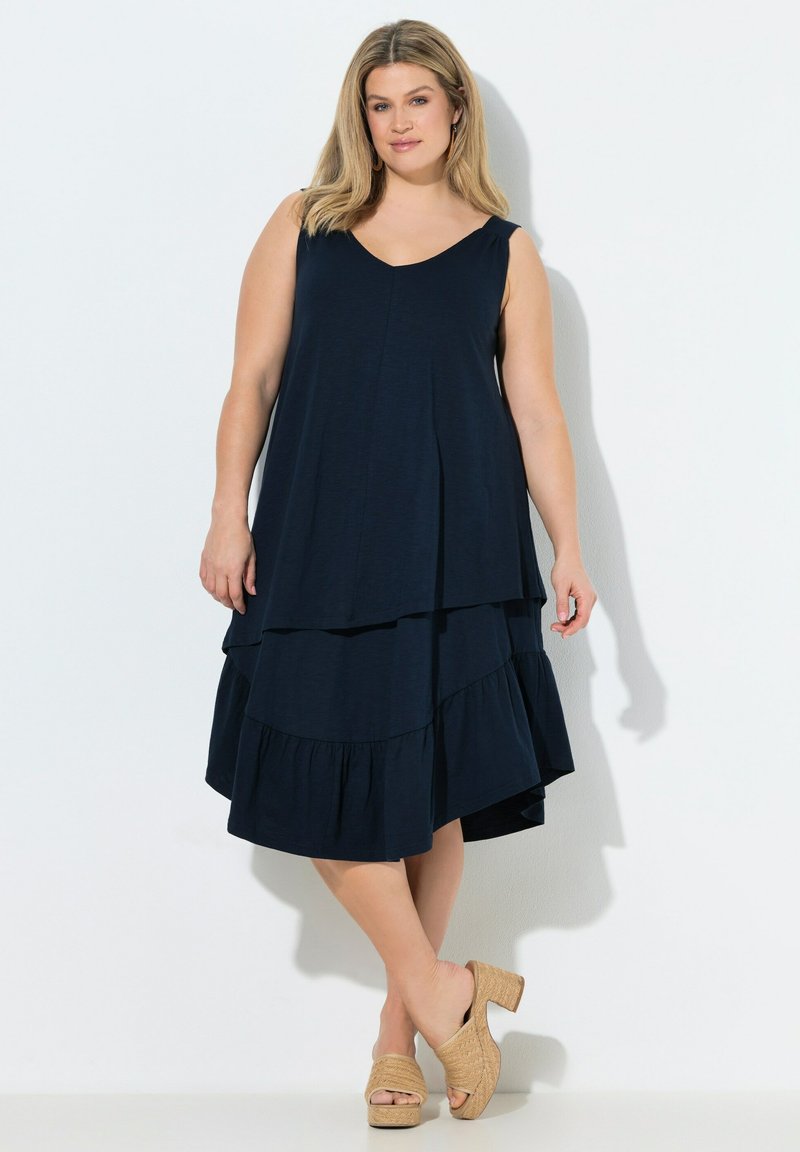 Femme portant une robe midi sans manches bleu marine à plusieurs couches et des sandales beiges tissées à talons, debout devant un fond blanc.