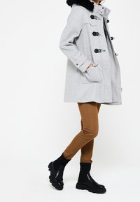 Šedý kabát z vlněné směsi s umělým kožíškem na límci, s černými háčkovými detaily a postranními kapsami. Spárováno s hnědými slim-fit kalhotami a černými kotníkovými botami.