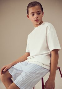 Pisana bela prevelika majica iz bombaža, v kombinaciji s svetlo modrimi kratkimi hlačami z lesenim vzorcem. Sedi na rdečem stolčku, osredotočena na priložnostna poletna oblačila.