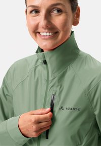 Giacca impermeabile verde chiaro con colletto alto. Presenta una tasca sul petto con zip e branding nero. Texture liscia e design aderente.