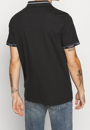 Mężczyzna w czarnej koszulce polo z krótkim rękawem z ozdobną taśmą Calvin Klein, niebieskich dżinsach i widocznymi tatuażami na obu ramionach, widziany od tyłu.