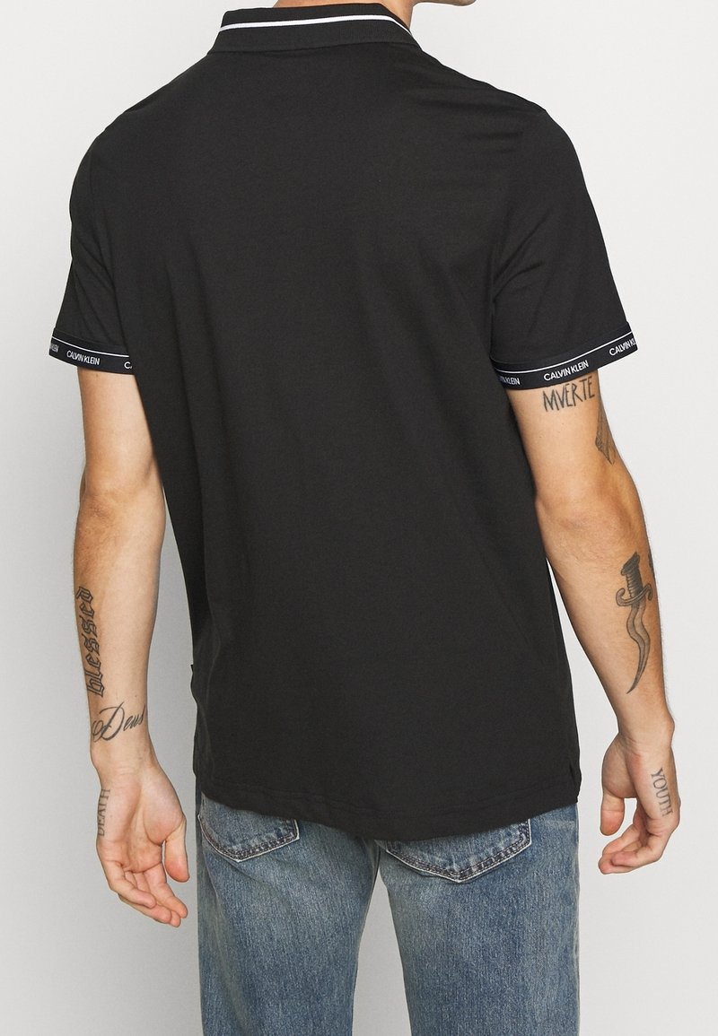Homme portant un polo noir à manches courtes avec bordure Calvin Klein, jean bleu et tatouages visibles sur les deux bras, vu de dos.