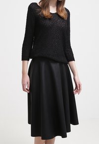 Vrouw in een zwarte gebreide trui met textuur en driekwartmouwen, met een hoogwaist zwarte midi-rok, staand tegen een effen achtergrond.