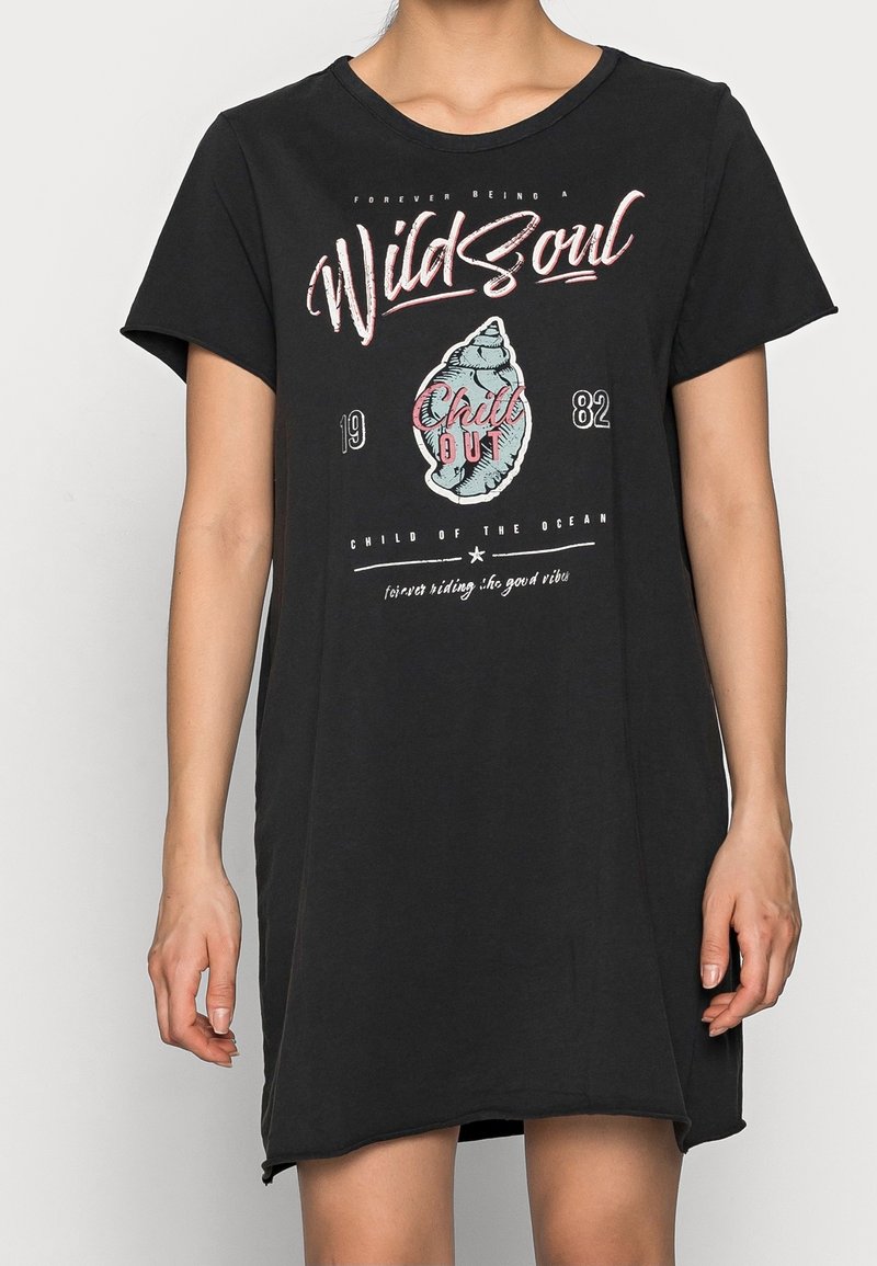 Femme portant une robe t-shirt noire oversize avec un visuel "Wild Soul" et coquillage, avec les textes "Child Out" et "Child of the Ocean."