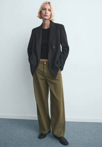 Blazer negro de doble botonadura sobre una camiseta negra ajustada, combinado con pantalones de pierna ancha en verde oliva. Textura suave, detalles mínimos de herrajes.