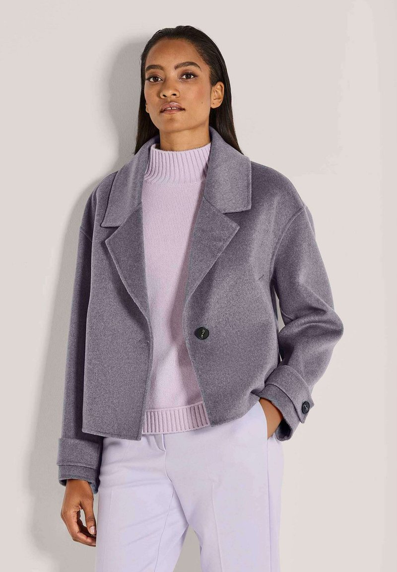 Eine Frau in einem helllila Rollkragenpullover, einer taillierten grauen Jacke mit schwarzen Knöpfen und helllila Hosen, steht mit einer Hand in der Tasche.