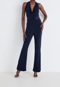 Marinblå jumpsuit med draperad hals, figurnära bodice, genomskinliga sidopaneler och utställda ben. Mjuk, elastisk tyg förhöjer komfort och rörelsefrihet.