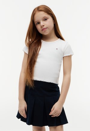 Νεαρή κοπέλα με μακριά κόκκινα μαλλιά, που φοράει λευκό κοντομάνικο πουκάμισο και σκούρο μπλε πιετέ φούστα, στέκεται μπροστά σε απλό φόντο.