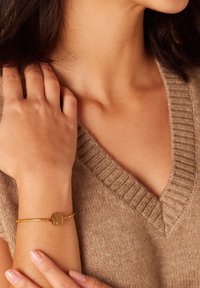 Gouden bangle armband met een ronde hanger met een boomontwerp. Draagbaar om de pols van een persoon in een tan kleurige, geribbelde trui. Huidtint is zichtbaar.