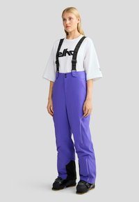 Lila wasserdichte Skihosen mit verstellbaren schwarzen Trägern, frontalen Reißverschlusstaschen und schwarzen Akzenten an den Bündchen, kombiniert mit einem weißen Grafik-T-Shirt.