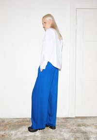 Witte blouse met lange mouwen, gecombineerd met blauwe wijde broek. De broek heeft een gladde textuur en zijzakken, aangevuld met zwarte schoenen.