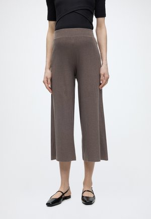 Γυναίκα με καφέ παντελόνι ψηλόμεσο, φαρδύ και cropped, και μαύρες μπαλαρίνες Mary Jane, στέκεται μπροστά σε απλό ανοιχτό φόντο.