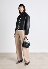 Giacca cropped in pelle nera sopra un dolcevita a righe bianche e nere, con pantaloni beige larghi. Tiene una piccola borsa nera con logo.