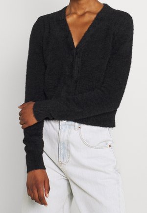 Osoba ubrana w czarny, puszysty, długi kardigan z dekoltem w serek, zestawiony z jasnoniebieskimi, wysokim stanem jeansami, na jednolitym tle.