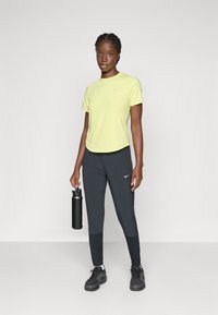 T-shirt Nike giallo a maniche corte, abbinato a pantaloni Nike grigio scuro. Il soggetto tiene in mano una bottiglia d'acqua nera, indossando scarpe da ginnastica nere.
