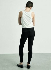 Černé strečové džíny skinny s vysokým pasem, zadními kapsami a hladkou texturou, na sobě má bílé tílko, pohled zezadu.
