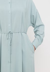 Ljusblå skjortklänning i mjukt tyg med dragsko i midjan, långa ärmar och sidofickor. Mjukt tyg och avslappnad passform.