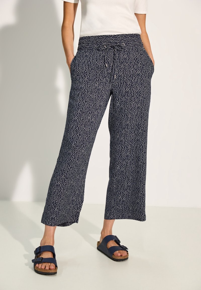 Pantalon large à motifs bleu marine avec taille smockée et poches latérales, associé à des sandales à glissière bleu foncé.