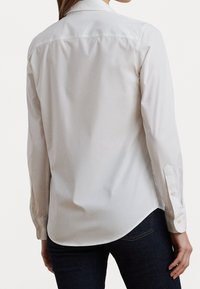 Kvinna sedd från ryggen, iförd en figursydd vit långärmad skjorta och mörkblå jeans mot en neutral ljus bakgrund.