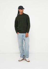 Donkergroen gebreid sweater, ruime pasvorm, ronde hals. Lichtblauwe losse spijkerbroek, witte sneakers met bruine zolen, zwart petje achterstevoren gedragen.