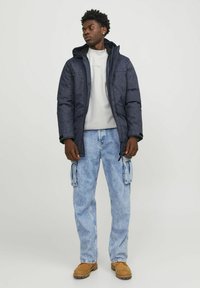 Chaqueta acolchada en azul marino con capucha, cierre de cremallera y bolsillos delanteros, combinada con jeans cargo en azul claro y botas marrones.