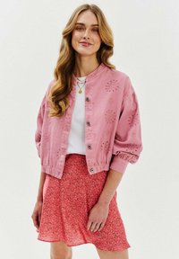 Różowa haftowana kurtka z okrągłym dekoltem i zapięciem na guziki, w połączeniu z różową spódnicą w kwiatowy wzór. Gładki materiał, luźny krój.