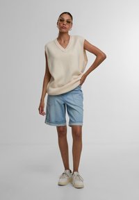 Débardeur crème sans manches avec un décolleté en V, associé à un short en denim bleu clair avec des ourlets. Des baskets blanches complètent le look.