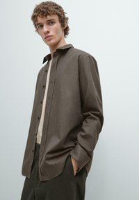 Brun tekstureret skjorte med knapper og spredt krave, båret over en lysefarvet top og kombineret med mørke bukser, poserende mod en neutral baggrund.