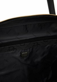 Wnętrze czarnej torby nylonowej z kieszonką na zamek, wytłoczonym logo "GUESS" oraz złotymi akcentami. Gładka tekstura z minimalnymi wzorami.