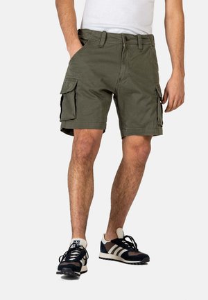 Mann trägt olivgrüne Cargo-Shorts mit Front- und Seitentaschen, weißes T-Shirt und schwarz-weiße Sneakers, steht vor weißem Hintergrund.