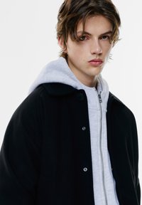 Schwarzer Außenmantel mit Kragen, versehen mit silbernen Druckknöpfen, über einem hellgrauen Reißverschluss-Hoodie getragen. Glatte Stofftexturen sind sichtbar.