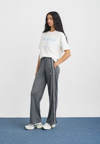 Szürke sportnadrág fehér oldalsávokkal, fehér pólóval, kék felirattal párosítva. Fehér és zöld sportcipővel viselve.