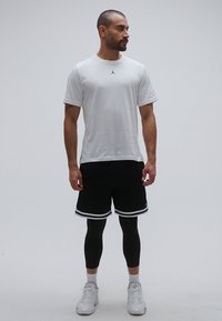 Λευκό πουκάμισο με κοντά μανίκια και μαύρο λογότυπο Jordan, μαύρο σορτς με λευκές ρίγες, μαύρο κολάν και λευκά αθλητικά παπούτσια.