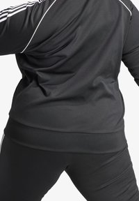 Černá sportovní bunda s bílými zvýrazňujícími pruhy, hladkou texturou, přiléhavým střihem a zaobleným spodním lemem, spárovaná s ladícími černými kalhotami.