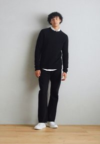 Black knit sweater over a white collared shirt, paired with black pants and white sneakers; model with curly hair posing against a neutral wall.