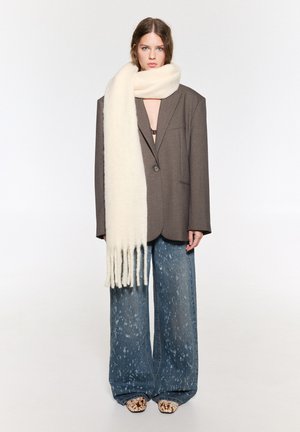 Blazer marrone oversize, sciarpa di lana crema con frange, jeans a gamba larga blu chiaro con macchie, e scarpe beige con motivo.