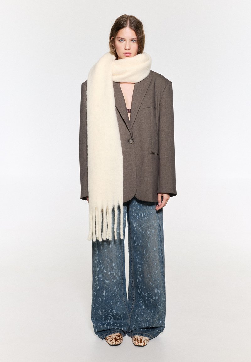 Överdimensionerad brun blazer, krämfärgad ullscarf med fransar, ljusblå prickiga vida jeans och mönstrade beige skor.