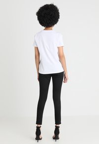 Kvinna med lockigt hår iförd en enkel vit t-shirt, svarta smala byxor och svarta högklackade sandaler, stående med ryggen mot kameran på en vit bakgrund.