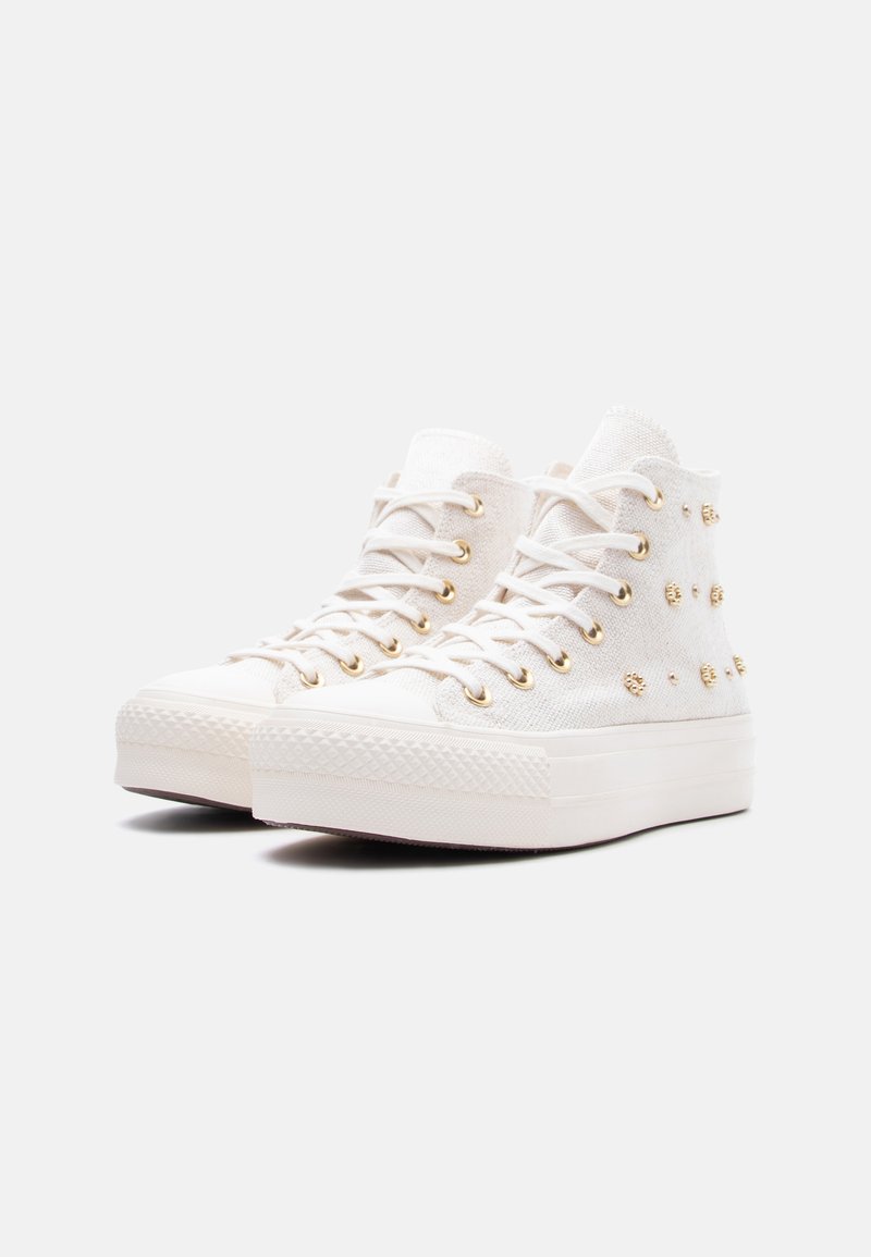 Scarpe da ginnastica alte bianche con suole platform, occhielli dorati e piccoli borchie dorate sulla tomaia in tessuto, mostrate leggermente inclinate su uno sfondo bianco.