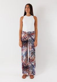 Top bianco senza maniche abbinato a pantaloni a gamba larga con una stampa di foglie di palma multicolore in blu, rosso e beige. Sandali marroni completano il look.