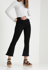 Mustat levenevät farkut, joissa on raakalaitaiset reunat, yhdistettynä istuvaan valkoiseen ribbineulottuun pitkähihainen paitaan ja mustiin korollisiin sandaaleihin nilkkahihnoilla.